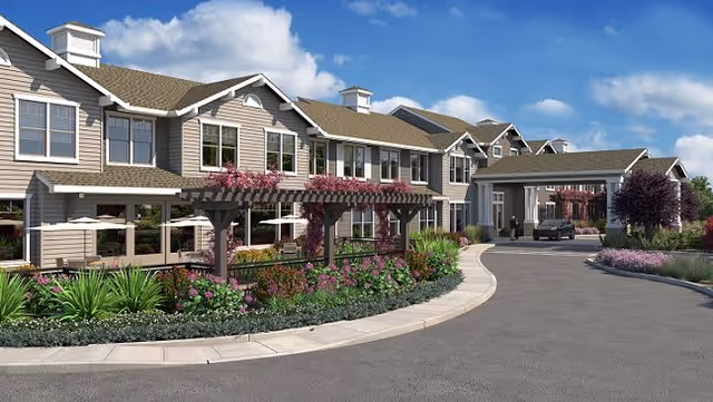 Exterior view of a senior living facility named Oakmont of Silver Creek, featuring a large two-story building with beige siding, multiple windows, and a covered entrance. The driveway curves in front of the building, surrounded by landscaped gardens with green shrubs, flowering plants, and a wooden pergola with climbing flowers. The sky is blue with scattered clouds.