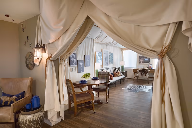 A cozy living room area viewed through an open curtain doorway. The room features a round wooden table with two chairs, a bench with patterned cushions, framed artwork on the walls, a desk with a chair, and a zebra-patterned rug on the wooden floor. Soft lighting comes from table lamps and a wall-mounted lantern.