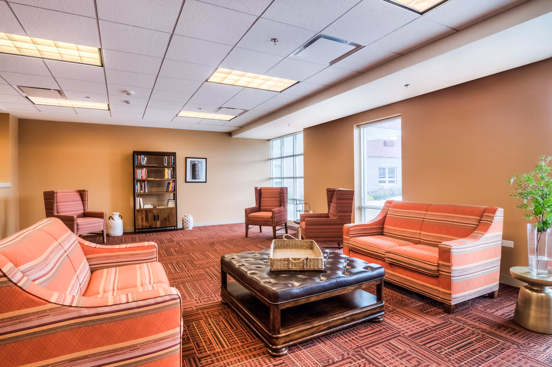 A spacious senior living facility lounge area with two striped sofas, four armchairs, a large square leather ottoman with a wicker basket on top, a bookshelf against the far wall, and large windows letting in natural light. The room has a warm color palette with beige walls and a patterned red carpet.