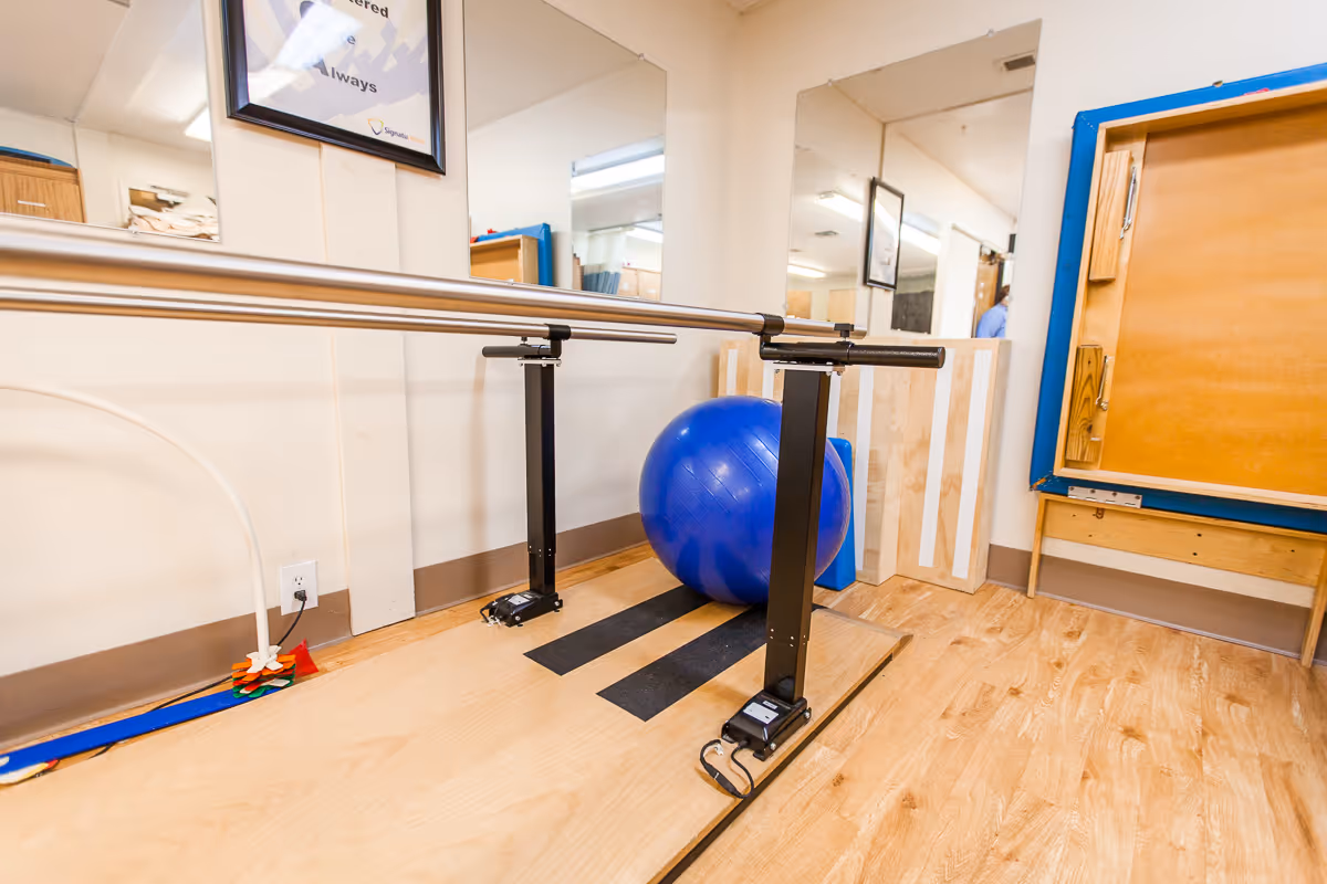 Physical therapy room with a wooden floor, parallel bars for walking exercises, a large blue exercise ball, mirrors on the wall, and some therapy equipment in the corner.