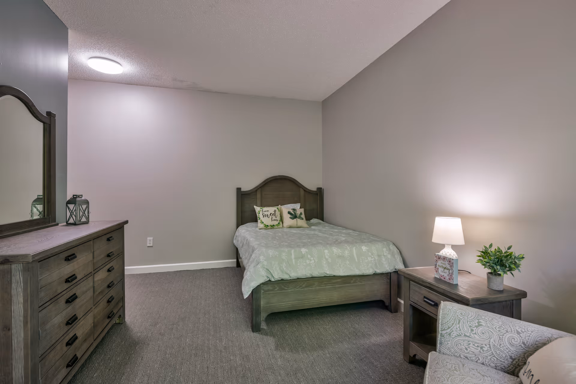 A simple bedroom with a wooden bed featuring a green patterned bedspread and two decorative pillows. To the left is a wooden dresser with a mirror and a lantern on top. On the right side of the bed is a wooden nightstand with a lamp, a small plant, and a decorative item. A patterned armchair is partially visible in the foreground. The walls are painted light gray and the floor is carpeted.