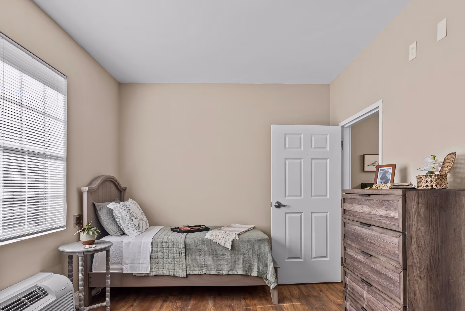 A simple bedroom with a single bed against a beige wall, covered with a green quilt and white sheets. There are two pillows on the bed, a small round wooden side table with a potted plant next to the bed, and a wooden dresser with a framed photo and a basket on top. A white door is partially open, and a window with blinds lets in natural light.