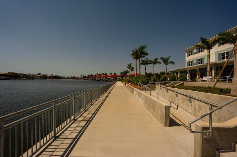 A wide waterfront promenade with metal railings, palm trees, red umbrellas in the distance, and a multi-story building on the right under a clear sky.