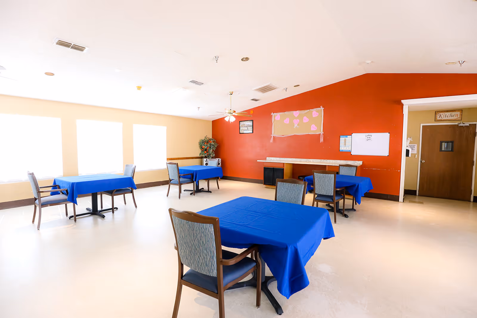 Bright dining room with tables draped in blue tablecloths, chairs, large windows, and a red accent wall.