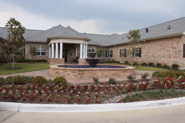Exterior view of a single-story brick building with a gray roof, featuring a circular fountain in front surrounded by a flower bed with red and white flowers. The building has a covered entrance with white columns and several windows.