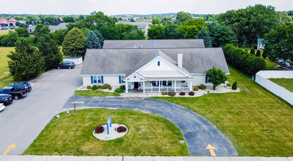 Aerial view of a single-story assisted living building with a curved driveway, front porch, lawn, and parked cars.