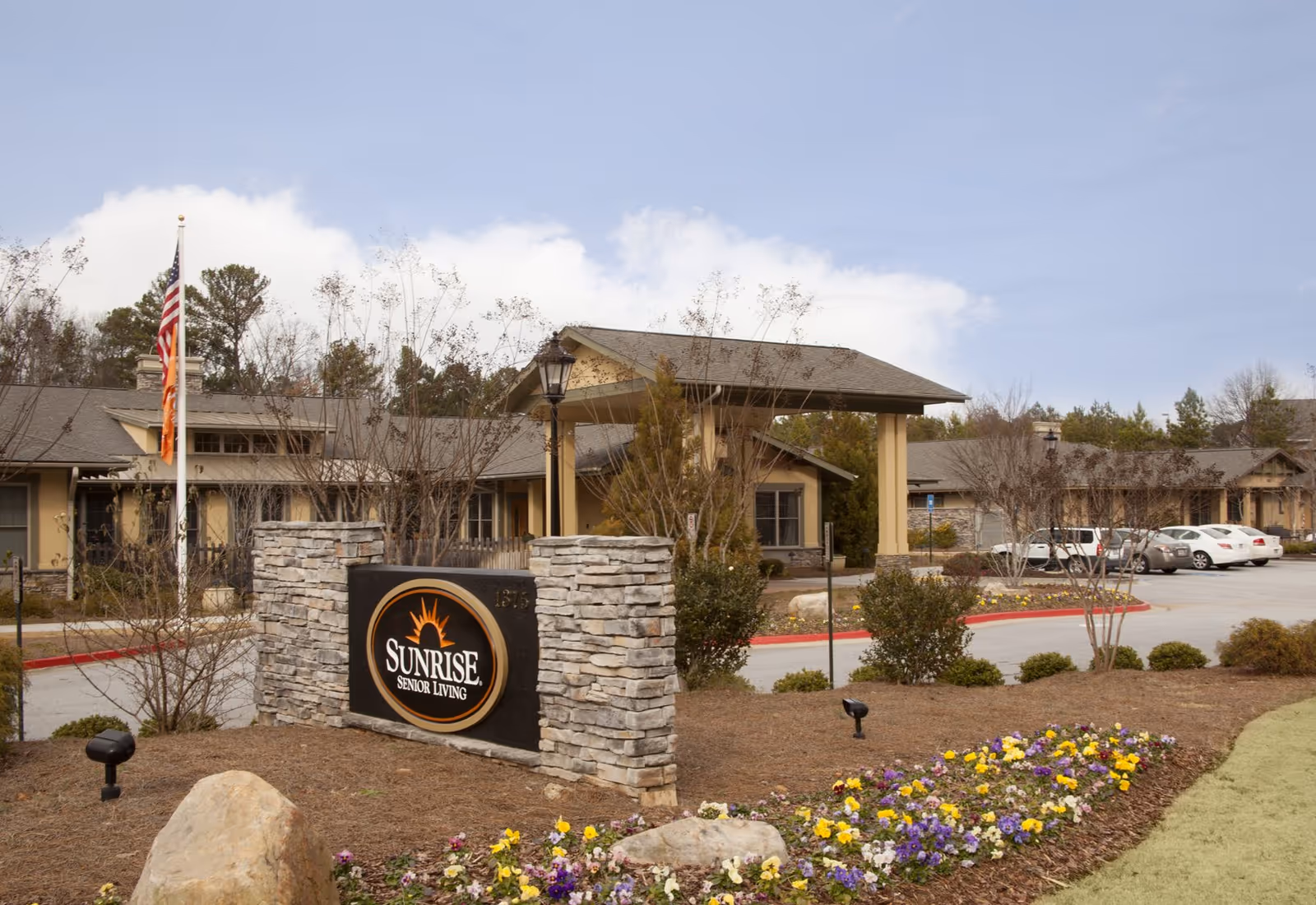 Exterior view of Sunrise Senior Living facility with a stone sign displaying the Sunrise logo in front. The building has a covered entrance, several windows, and a parking area with cars. There are flower beds and landscaping around the sign and entrance.