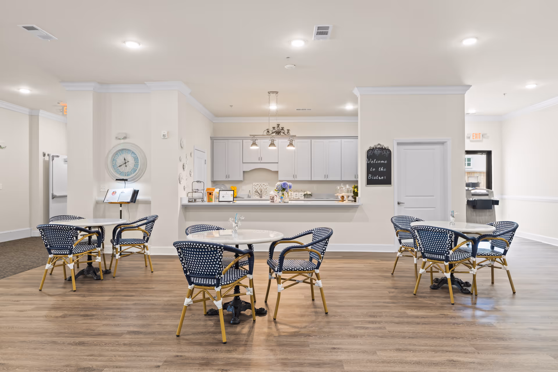 Communal dining area with round tables and blue-patterned bistro chairs in front of a light-colored kitchenette and serving counter.