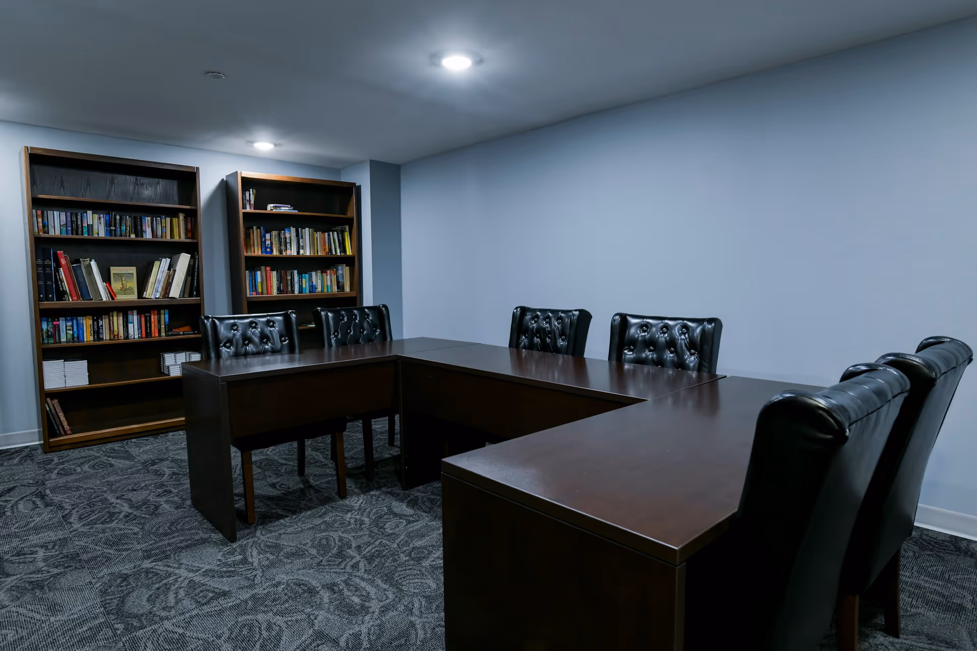 A quiet room with dark wooden tables arranged in a U-shape and black leather tufted chairs around them. Two wooden bookshelves filled with books are against the wall. The room has gray patterned carpet and light gray walls with ceiling lights.