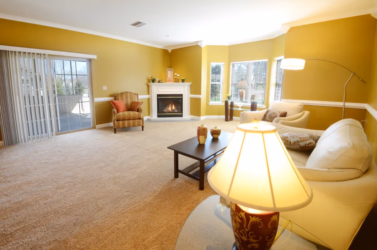 A bright living room with beige carpet, yellow walls, and white trim. The room features a white fireplace with decorative items on the mantel, a striped armchair with red cushions, a white sofa and armchair with patterned pillows, a dark wooden coffee table with decorative vases, a floor lamp, a table lamp in the foreground, and large windows letting in natural light. Sliding glass doors lead to an outdoor patio.