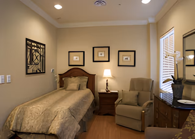 A cozy senior bedroom with a single bed covered in beige bedding, a wooden headboard, and a matching pillow. Next to the bed is a wooden nightstand with a lit table lamp. On the right side of the room, there is a comfortable armchair with a cushion, a wooden dresser with a large mirror, and a potted plant. The walls are decorated with three framed pictures above the bed and one framed artwork on the left wall. A window with blinds allows natural light into the room.