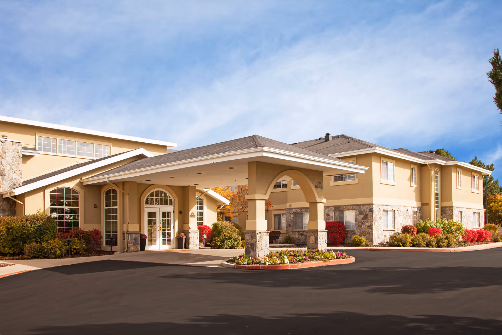 Exterior view of Chancellor Gardens senior living facility showing a beige two-story building with stone accents, large windows, and a covered entrance with pillars. The driveway and landscaped flower beds with colorful bushes are visible under a partly cloudy blue sky.