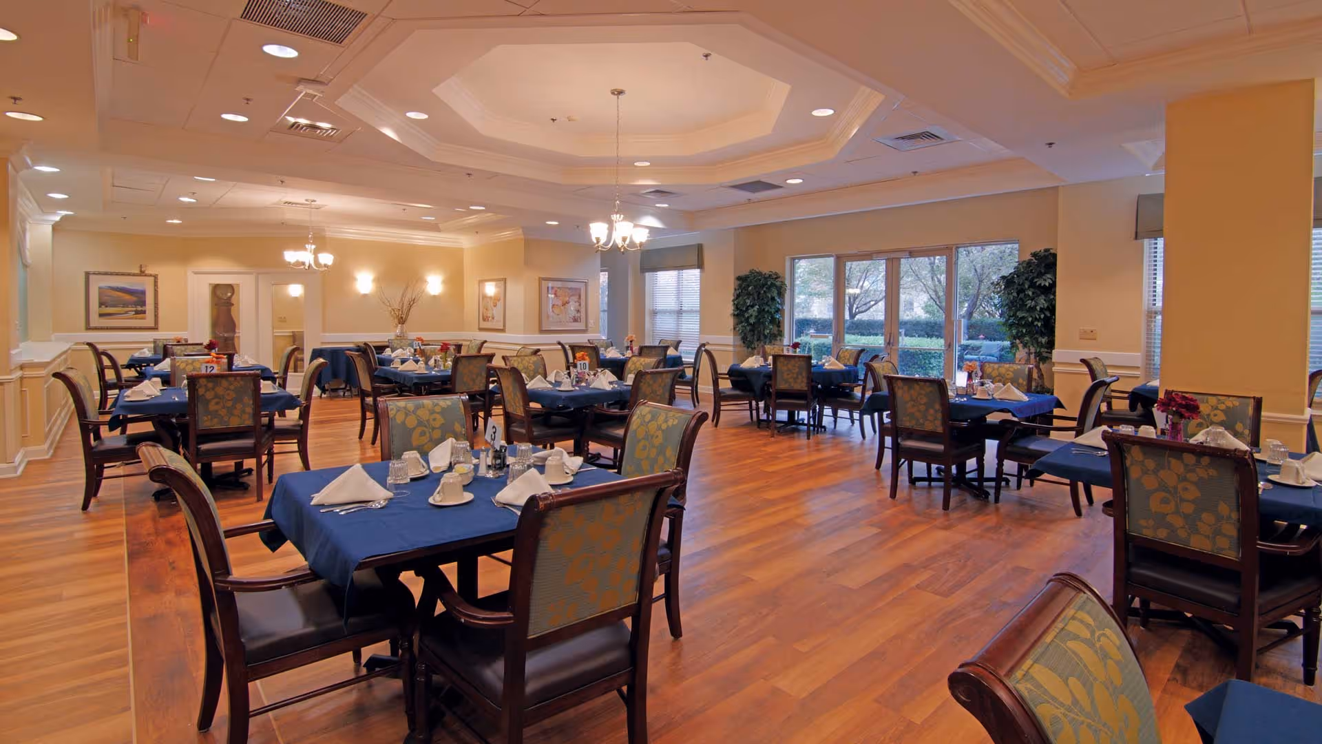 Dining room with multiple set tables covered in blue tablecloths, wooden chairs, chandeliers, and large windows overlooking landscaping.