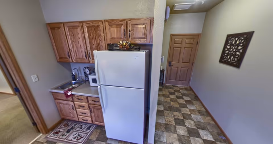 Small kitchen area with wooden cabinets, a white refrigerator, a microwave, and a sink. The floor has a checkered tile pattern. Adjacent to the kitchen is a hallway with a wooden door and a decorative wall hanging.
