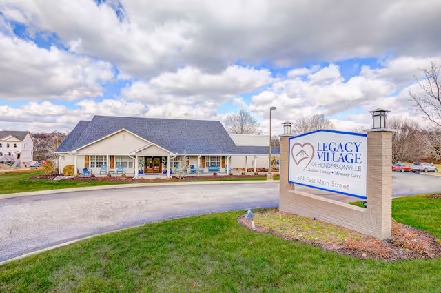 Exterior view of Legacy Village of Hendersonville, showing a single-story building with a dark roof and a covered entrance. In the foreground, there is a large sign with the facility's name and address, surrounded by green grass and a curved driveway. The sky is partly cloudy.