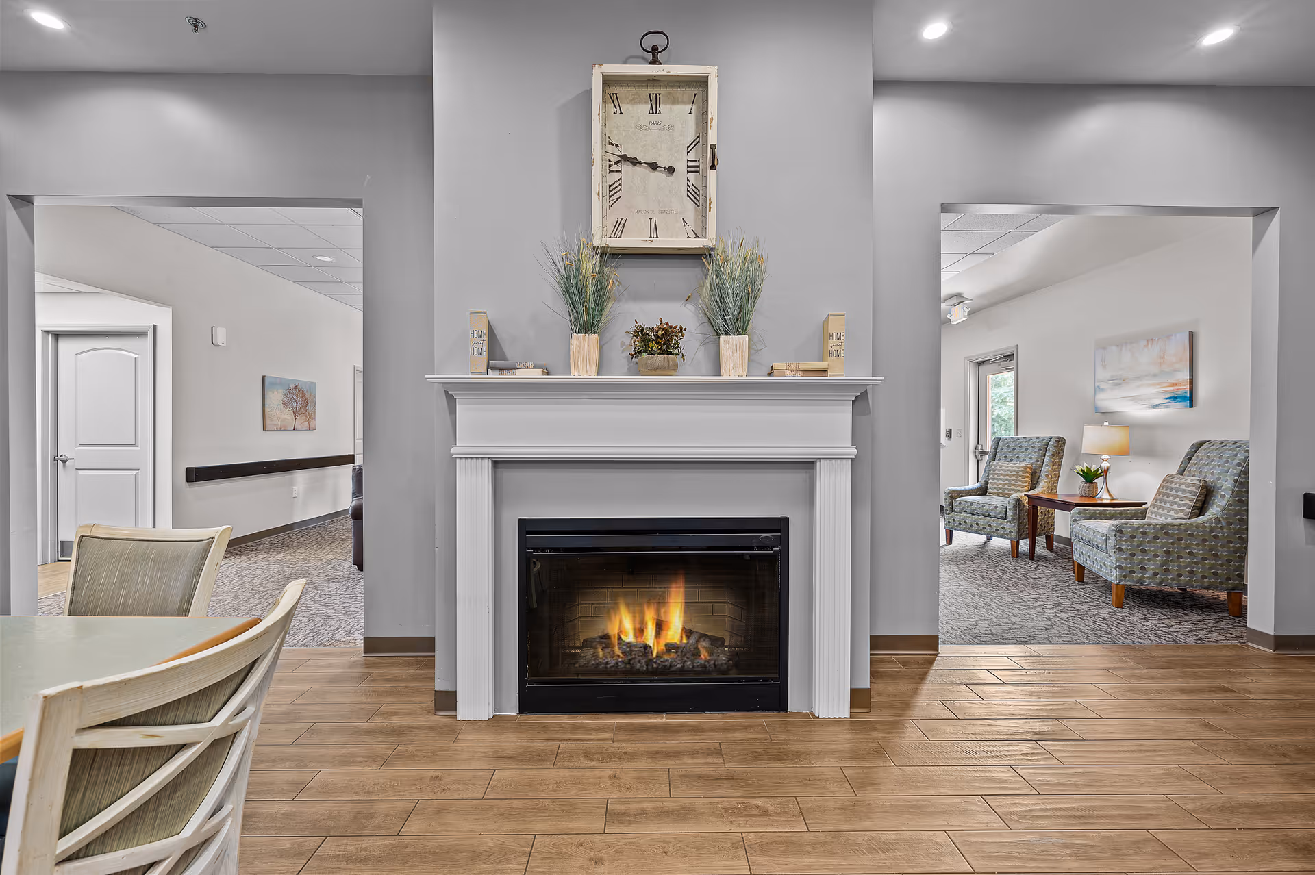 Interior view of a senior living facility featuring a lit fireplace with a mantel decorated with plants, books, and a large clock above it. On the left, there is a dining area with chairs and a table, and on the right, a sitting area with two upholstered armchairs, a side table with a lamp, and a painting on the wall.