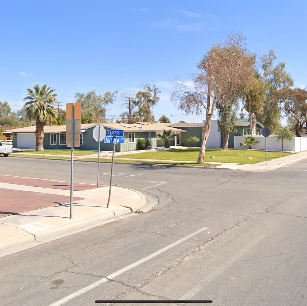 Street view of a single-story residential building with green siding and a beige roof at the corner of Cypress Drive and Aurora Drive. The scene includes a palm tree, other trees, a well-maintained lawn, and clear blue sky.