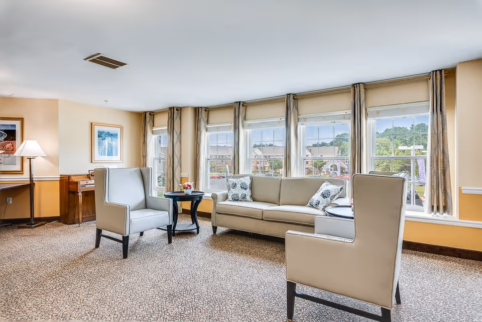 Bright and spacious living room with large windows letting in natural light, featuring a beige sofa with patterned cushions, two beige armchairs, a small round black table with a flower vase, a floor lamp, and framed artwork on the walls.