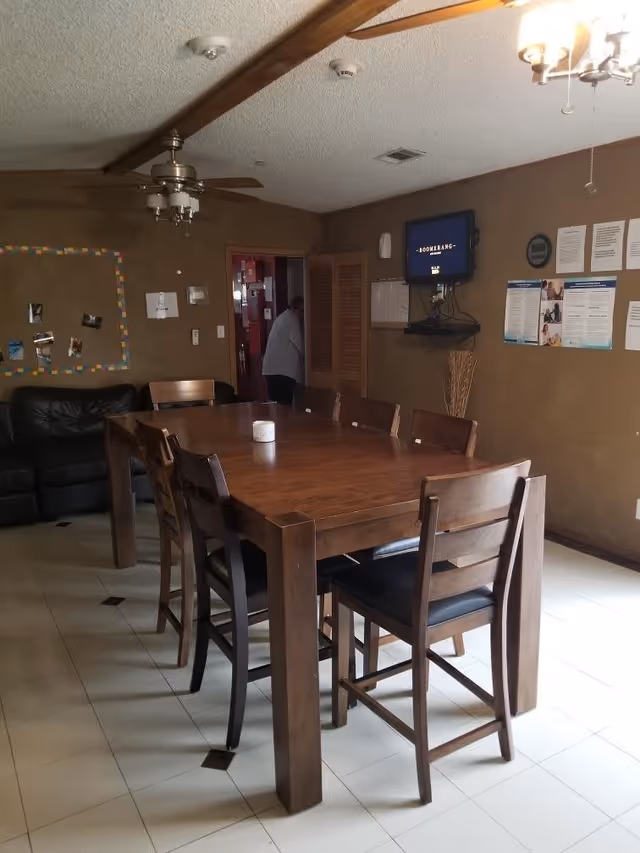 A dining room with a large wooden table surrounded by eight wooden chairs with black cushions. There is a black leather couch against the left wall, a ceiling fan with lights above the table, and a wall-mounted TV on the right wall. Various papers and notices are pinned on the wall near the TV. A person is partially visible in the doorway at the back of the room.
