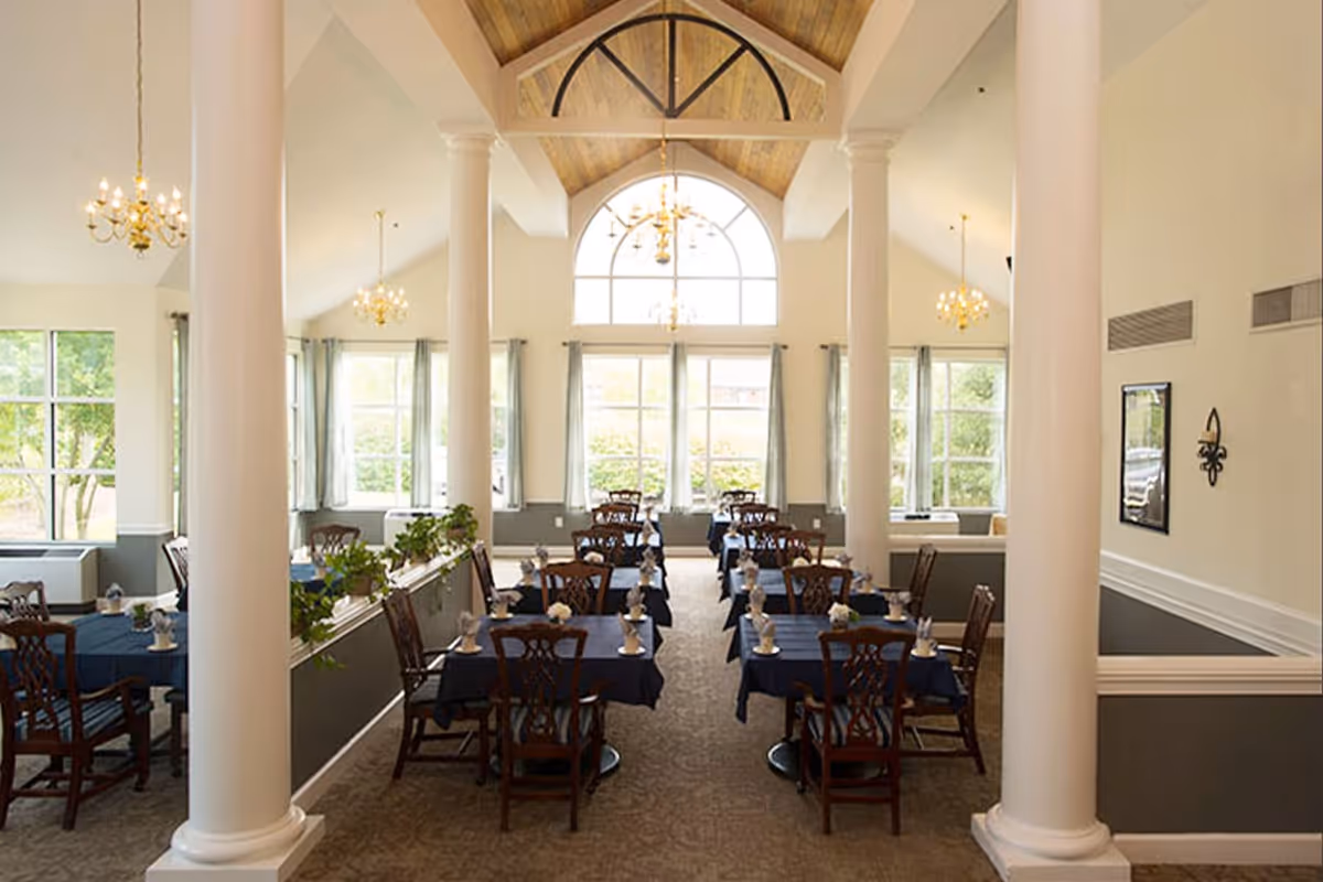 A spacious dining room with high vaulted ceilings and large arched windows allowing natural light to fill the room. The room features multiple tables covered with dark blue tablecloths, each set with napkins and small floral centerpieces. Elegant chandeliers hang from the ceiling, and white columns line the center of the room.