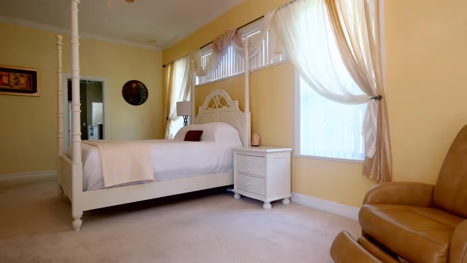 A bright bedroom with a white four-poster bed covered in white bedding and a small dark pillow. Next to the bed is a white nightstand with a lamp and a small decorative item. The room has light yellow walls, beige carpet, and large windows with sheer curtains letting in natural light. A brown leather armchair is positioned near the window. There is a doorway leading to another room visible in the background.