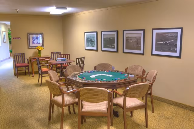 A well-lit interior room with a large octagonal poker table surrounded by eight cushioned chairs. The poker table has playing cards and poker chips arranged on it. On the wall behind the table are four framed black and white photographs of airplanes. In the background, there is a small table with a vase of yellow flowers and two chairs. The room has beige walls and carpeted flooring.
