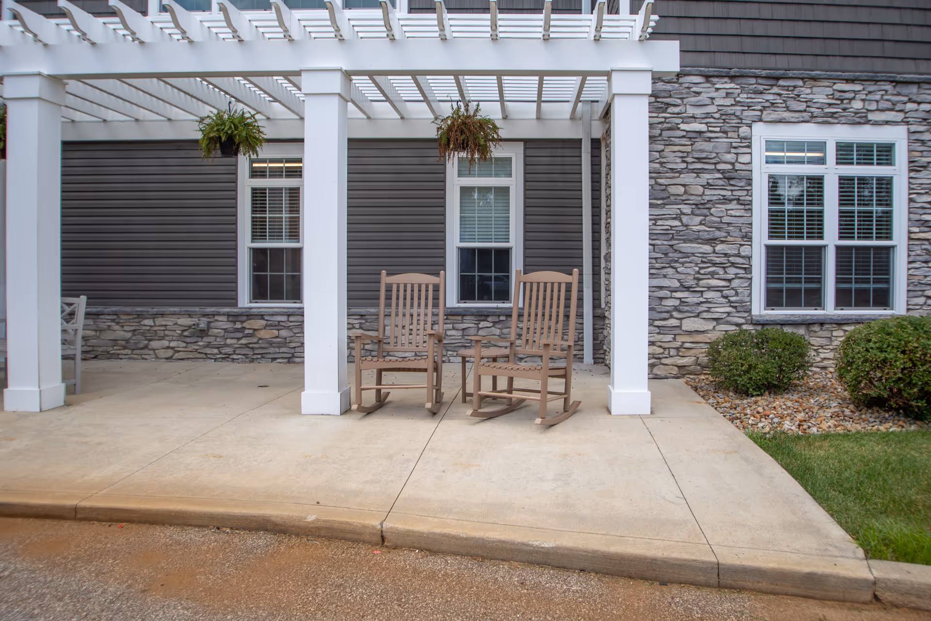 Outdoor patio area with two wooden rocking chairs placed under a white pergola attached to a building with stone and gray siding. There are two hanging plants above the chairs and windows with white frames and blinds on the building wall.