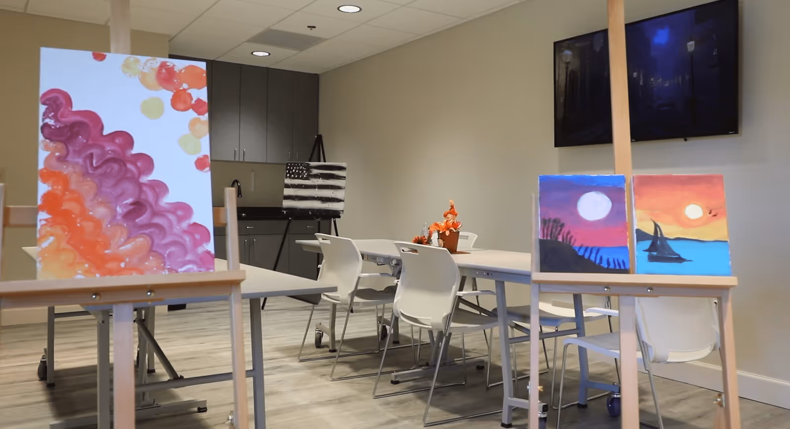 Interior activity room with easels displaying colorful paintings, tables and white chairs, cabinets and a wall-mounted TV.
