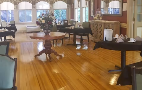 A spacious dining room with polished wooden floors, several tables covered with black tablecloths, chairs, a round wooden table with a large floral arrangement in the center, large windows with white curtains, and a wooden sideboard with decorative items and a mirror above it.