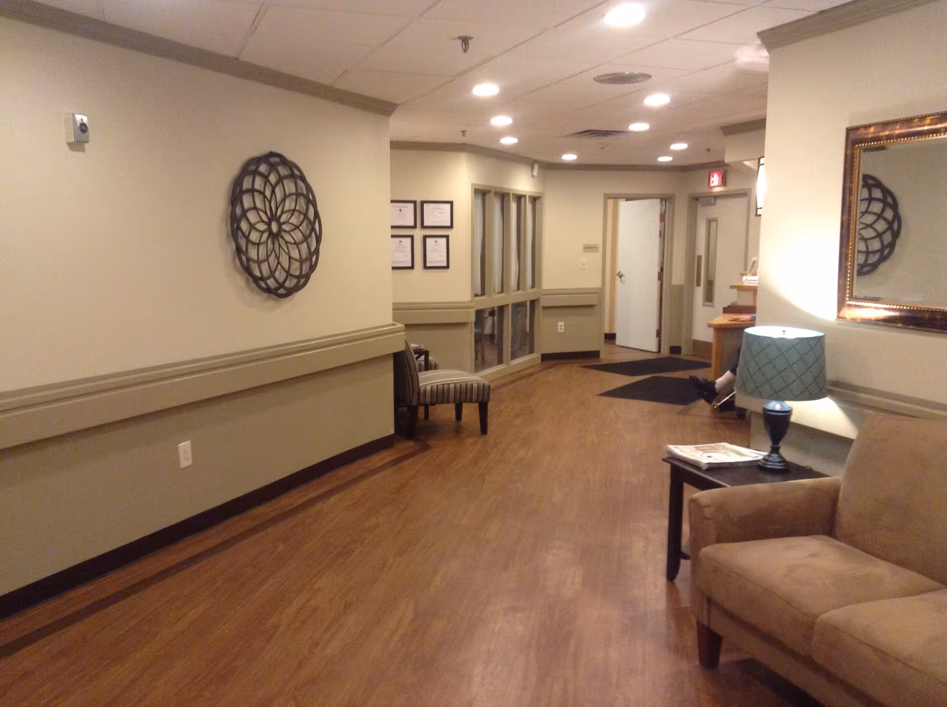 Interior hallway of a senior living facility with wood flooring, beige walls, a brown couch, a side table with a lamp, a decorative wall piece, and framed certificates on the wall. There is a chair near the end of the hallway and an open door leading to another room.