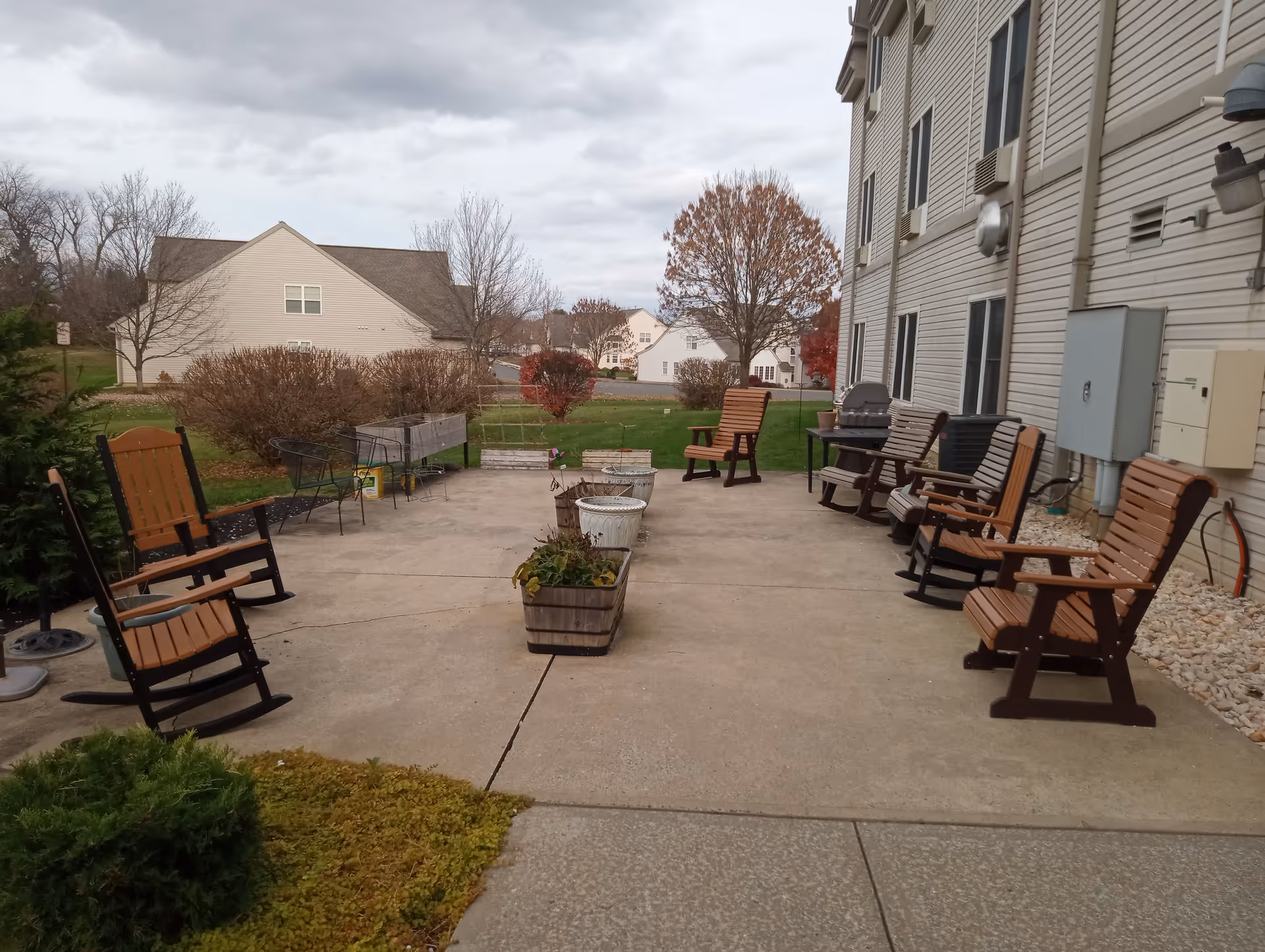 Outdoor patio area with several wooden rocking chairs and regular chairs arranged along the sides. There are planter boxes with plants in the center of the concrete patio. The patio is adjacent to a building with beige siding and windows. In the background, there are trees with autumn foliage and neighboring houses under a cloudy sky.