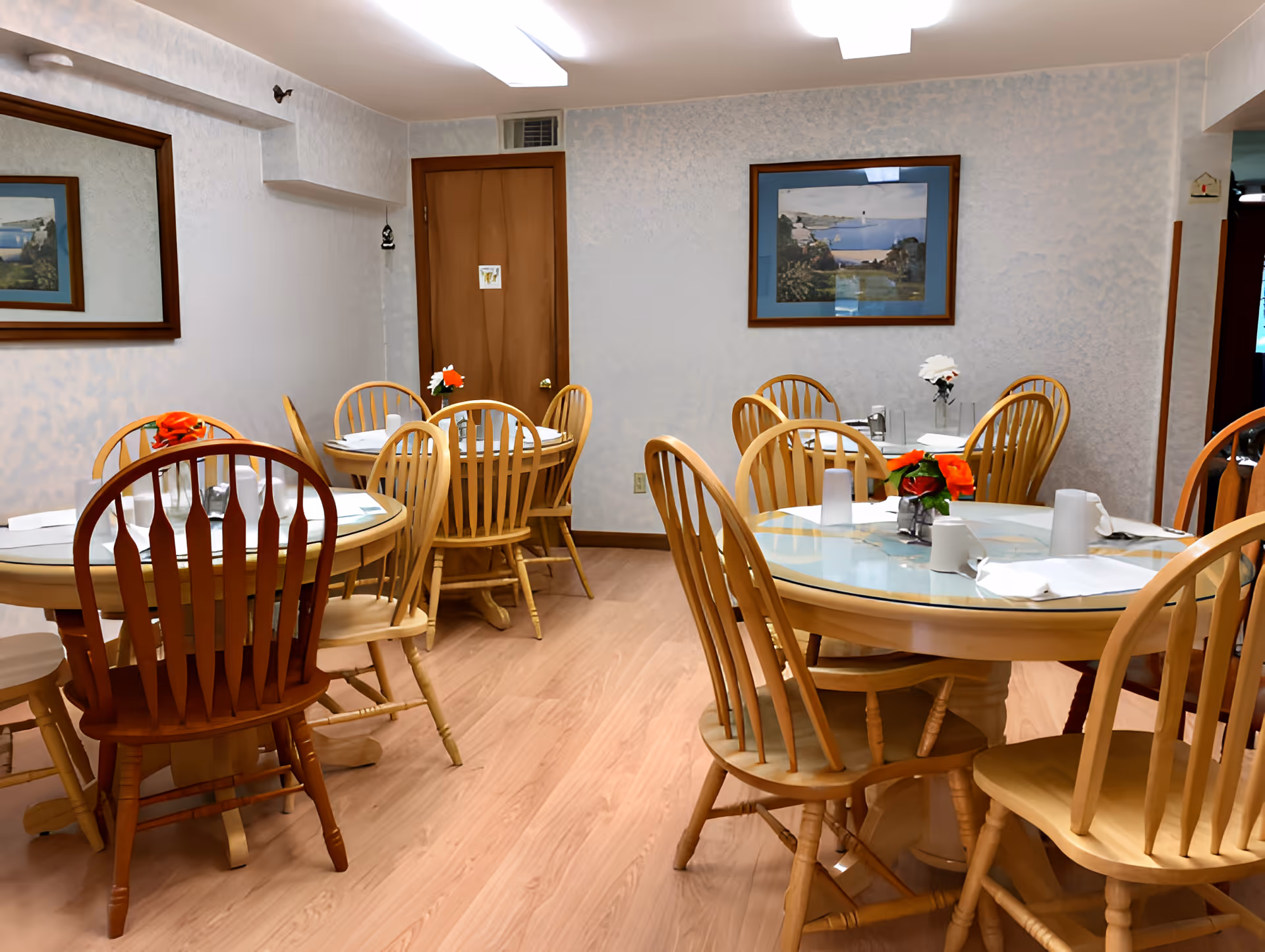 Small dining room with round tables set with place settings and flower centerpieces, surrounded by wooden chairs and framed artwork on the walls.