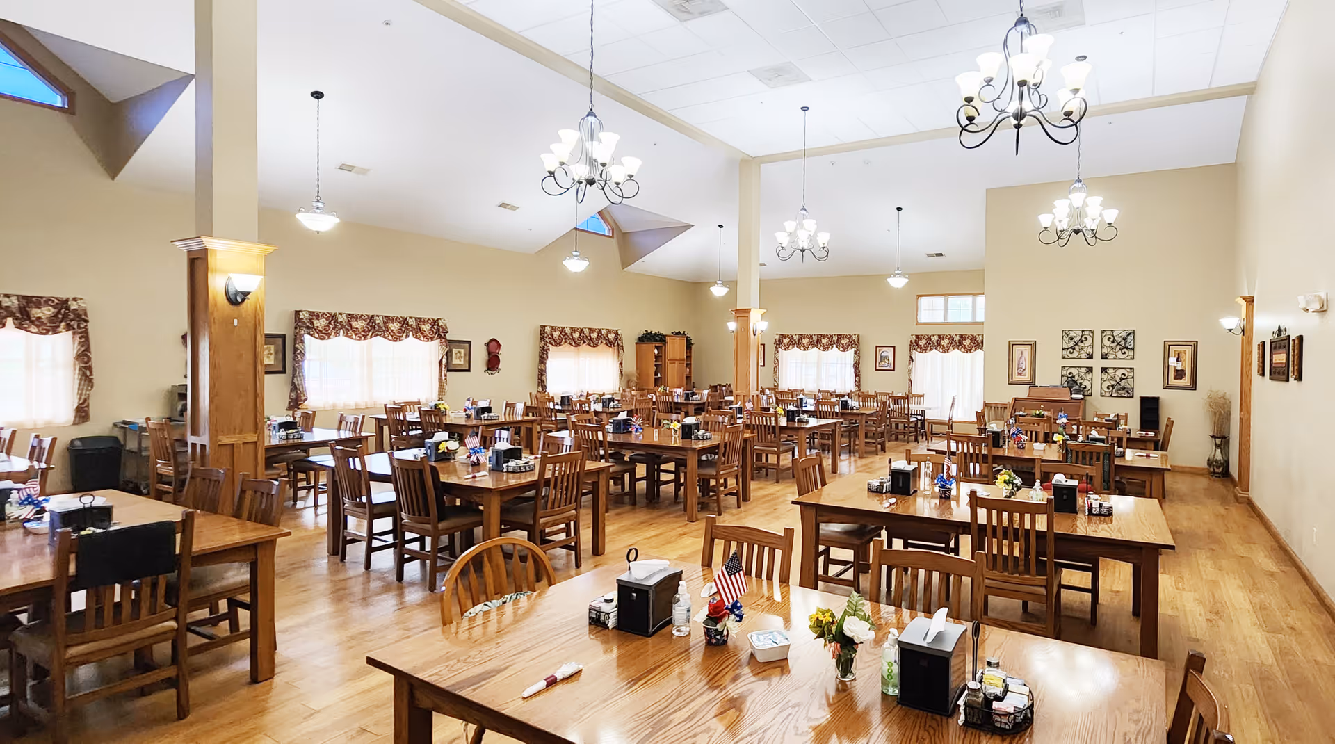 Large, bright communal dining room with wooden tables and chairs, chandeliers, and place settings.