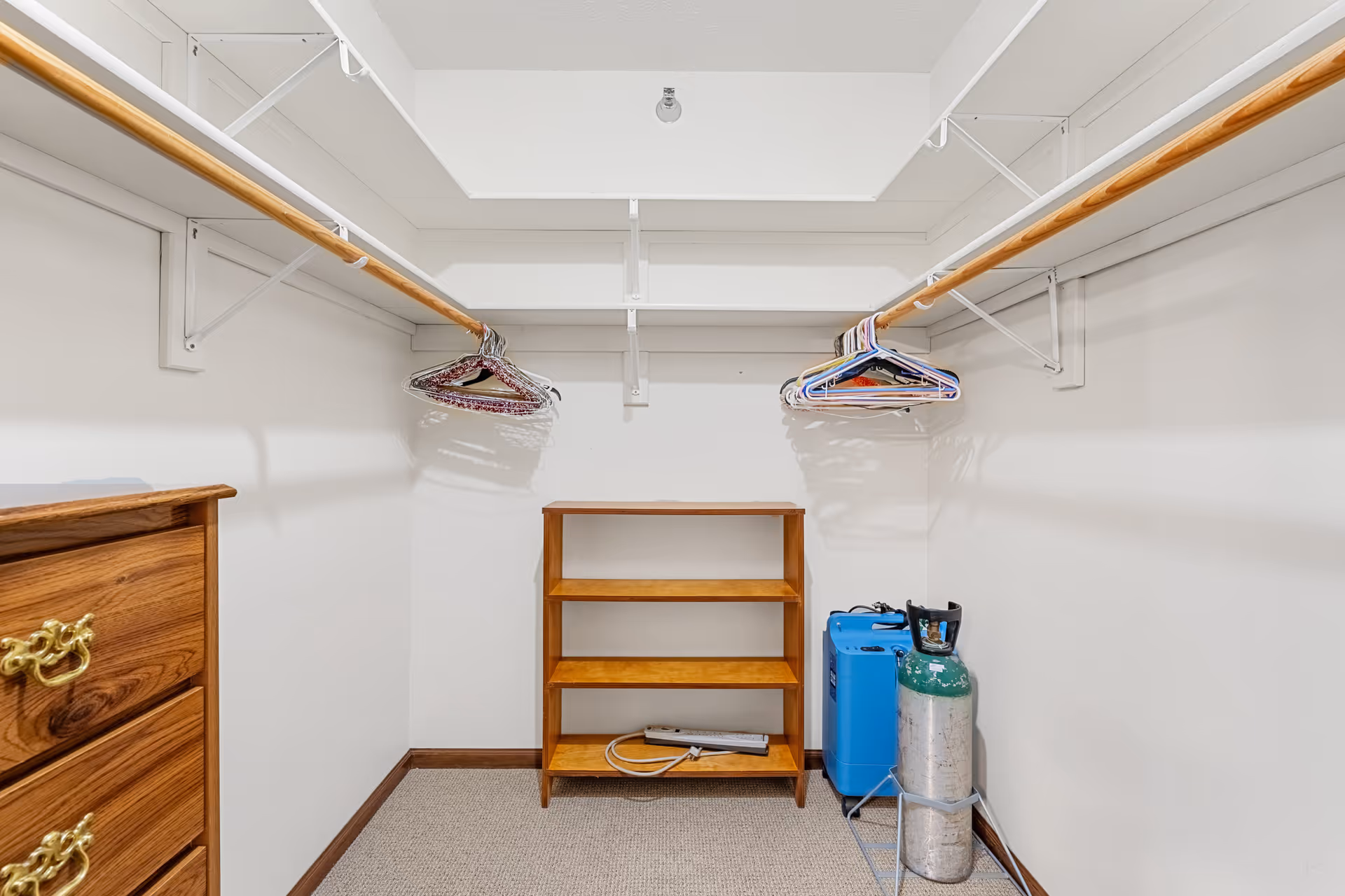 A walk-in closet with wooden shelves and hanging rods on both sides. There are several empty hangers on the rods, a wooden dresser on the left, and an oxygen tank and a blue device on the floor to the right.