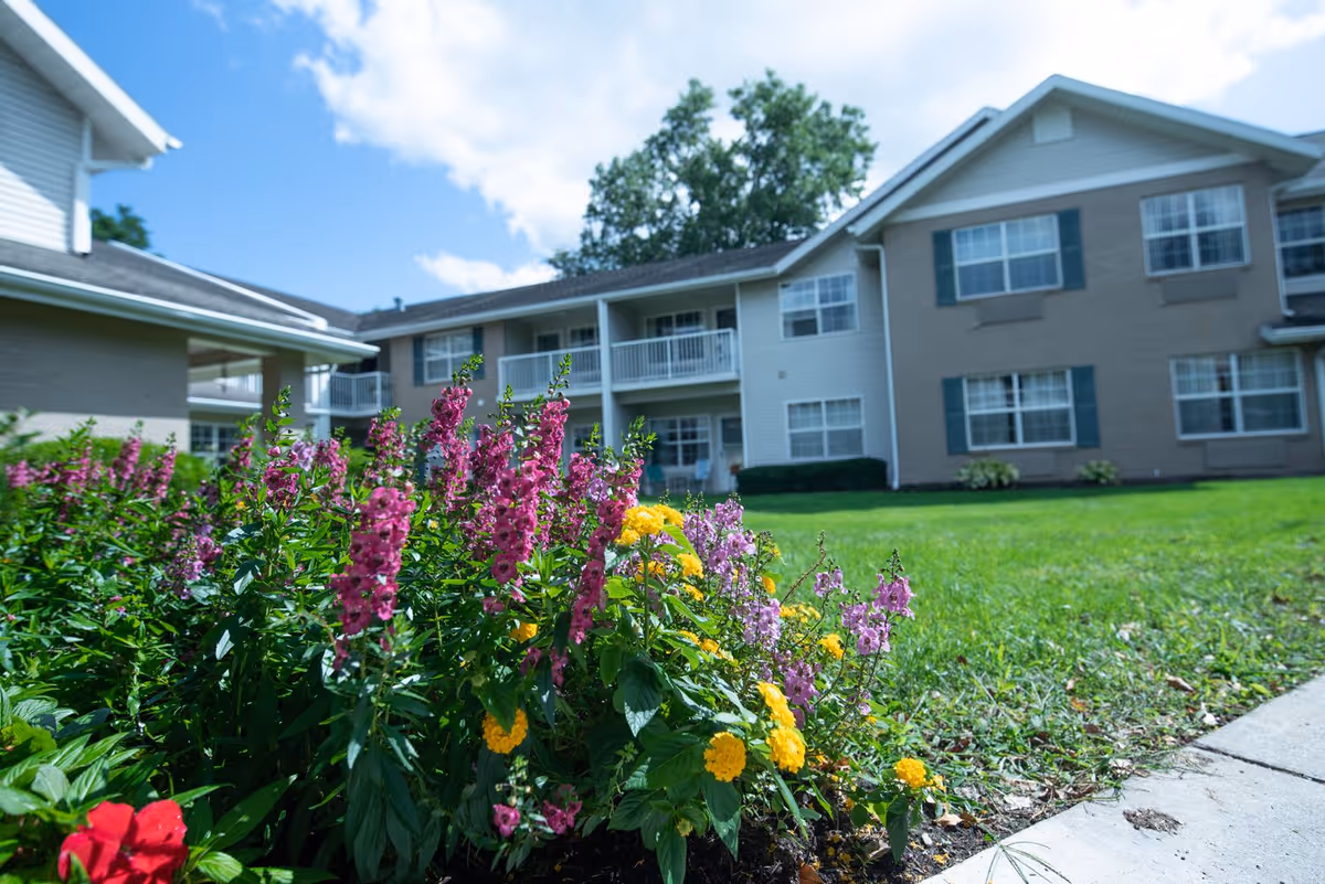 Colorful flowers including pink, yellow, and red blooms in a garden bed with a green lawn and a two-story senior living building in the background under a partly cloudy blue sky.