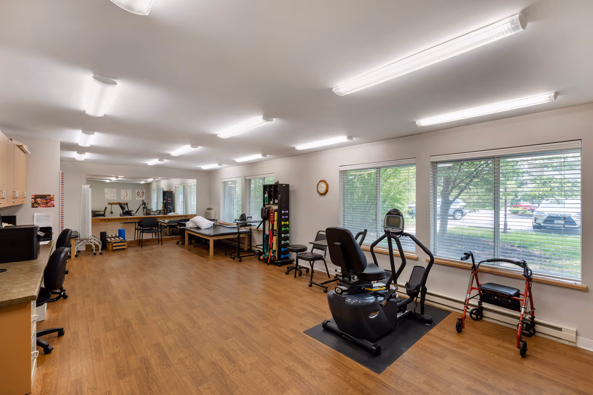 A spacious senior living facility exercise and therapy room with wooden flooring, large windows with blinds, exercise equipment including a recumbent bike, a walker, therapy tables, chairs, and various physical therapy tools. The room is well-lit with ceiling lights and has a clean, organized appearance.