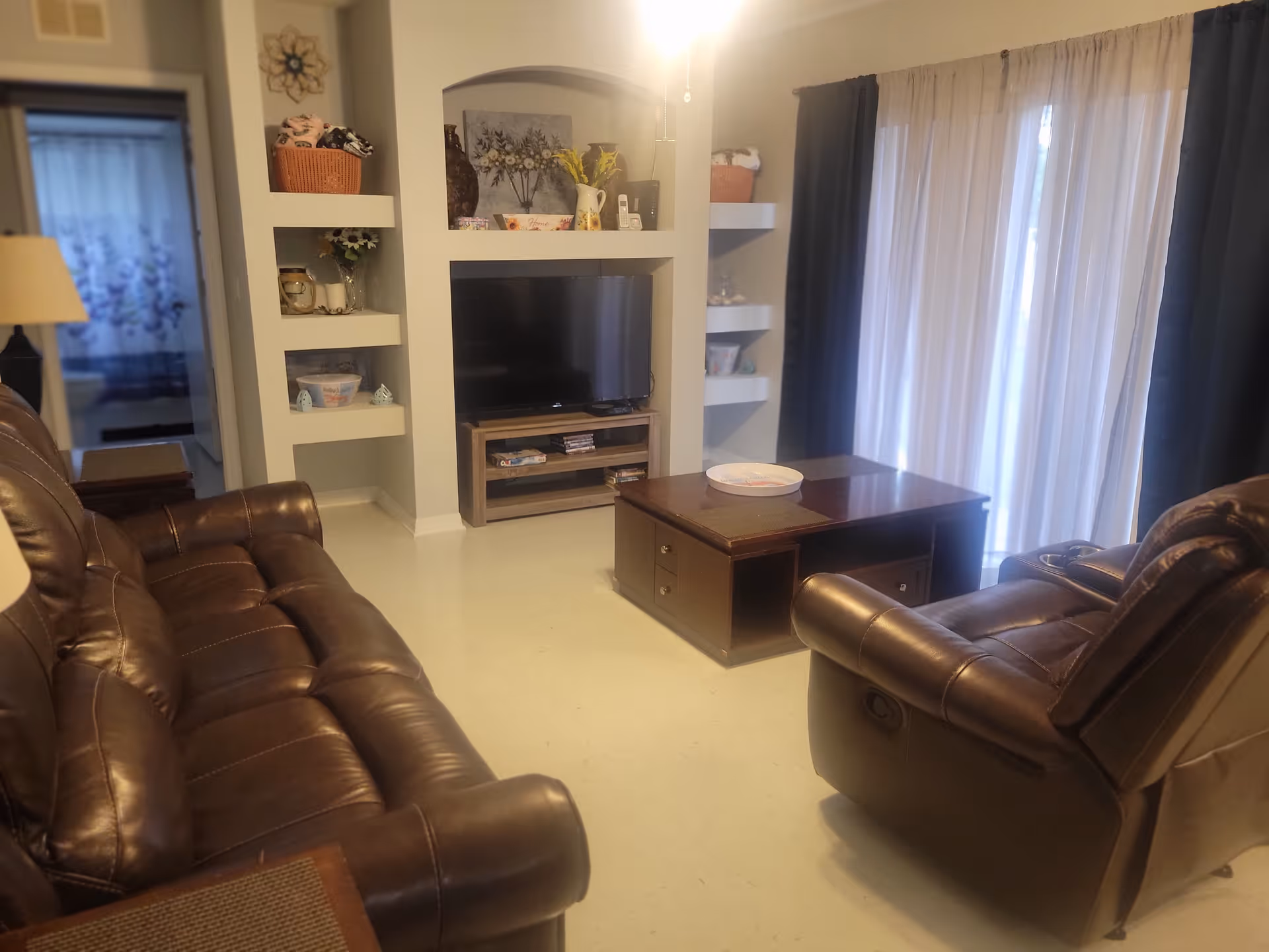 A cozy living room with two brown leather recliners and a matching sofa facing a flat-screen TV mounted in a white built-in wall unit with shelves holding decorative items. A wooden coffee table with a tray is in the center, and large windows with sheer and dark curtains allow natural light to enter the room.
