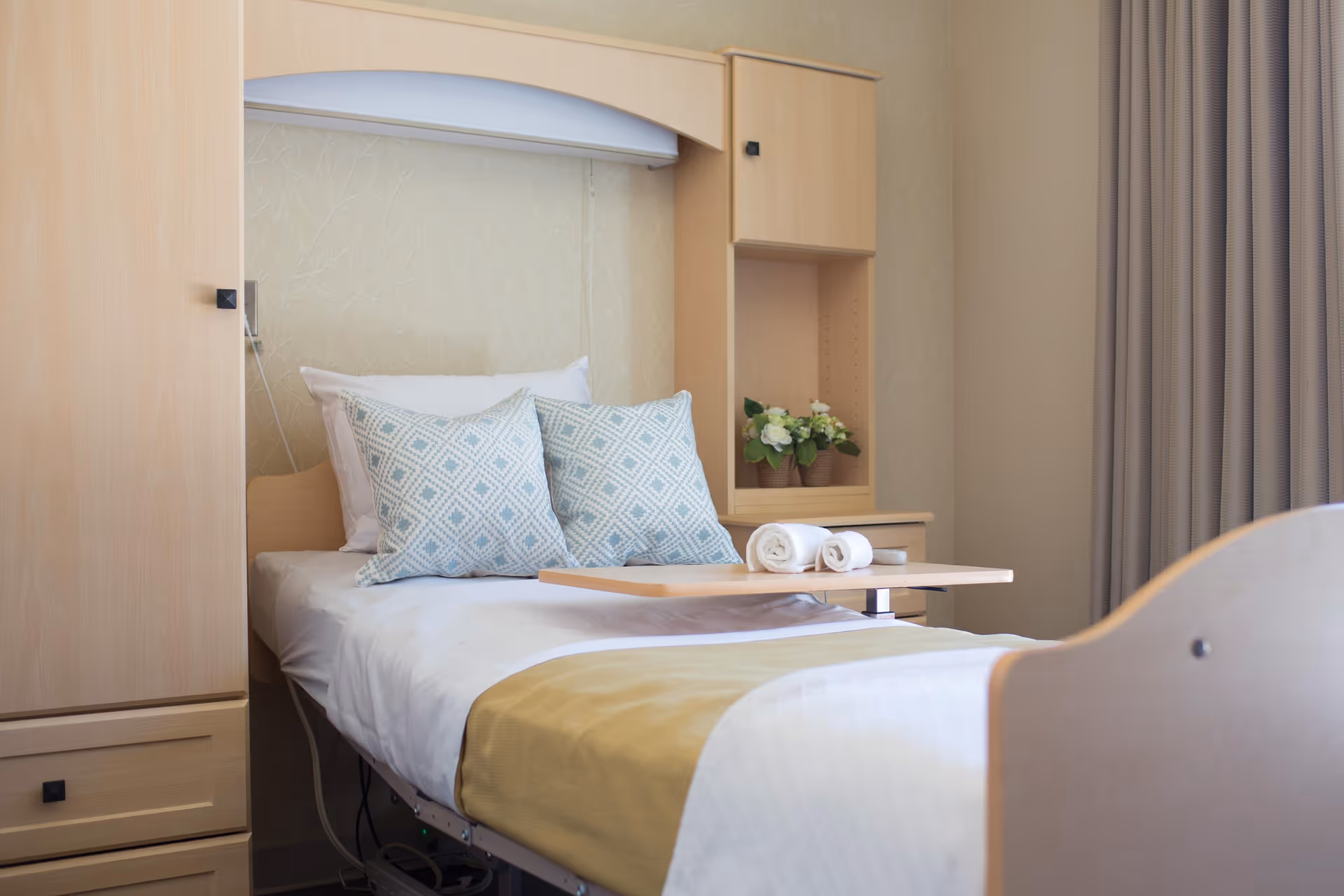 A neatly made single bed in a senior living facility room with two patterned pillows, a yellow bed runner, and a wooden overbed table holding two rolled white towels. The bed is surrounded by light wood cabinetry and a small shelf with a potted plant. Beige curtains cover the window on the right side.