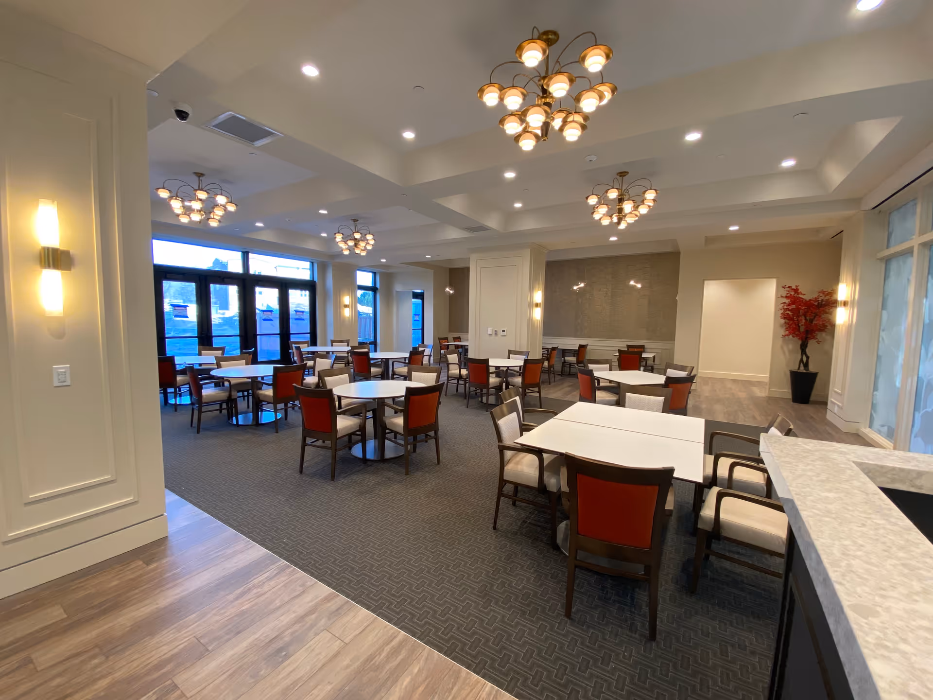 A spacious dining room with multiple round and rectangular tables surrounded by chairs with red and beige upholstery. The room features modern chandeliers hanging from a white ceiling with recessed lighting. Large windows and glass doors allow natural light to enter, and a decorative red plant is placed in the corner near a doorway. The floor has a combination of carpet and wood finishes.