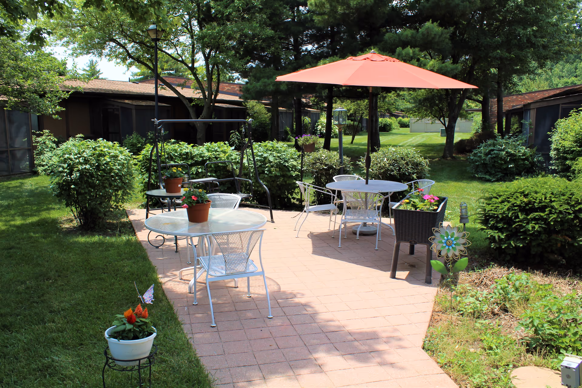 Outdoor patio area at Claremont Retirement Village featuring two round metal tables with chairs, one table shaded by a large orange umbrella. There are potted plants on the tables and surrounding greenery including bushes and trees. A swing bench is visible in the background along with a well-maintained lawn and residential buildings.