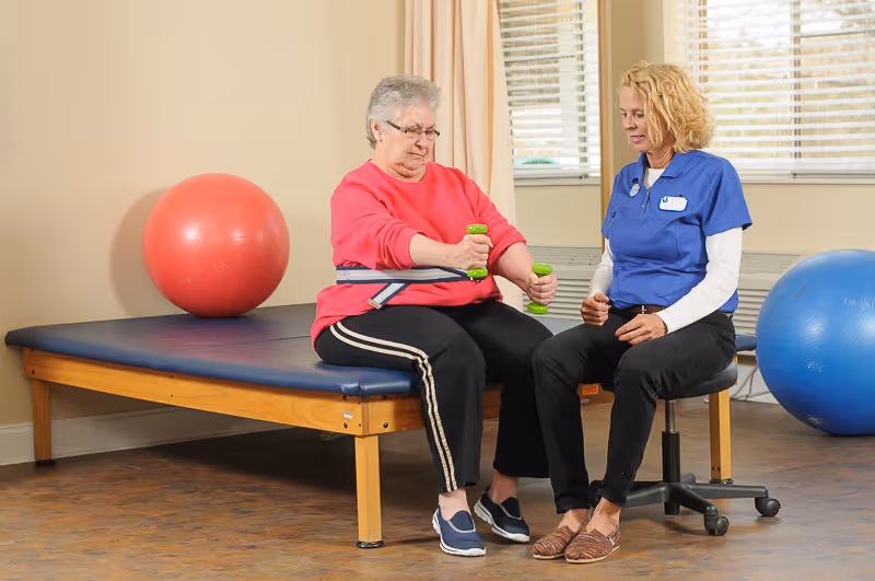 An elderly woman in a red sweater and black pants is seated on a padded exercise table holding small green dumbbells, while a female healthcare worker in a blue uniform sits on a rolling stool next to her, providing assistance. There are two large exercise balls, one red and one blue, in the room with beige walls and windows with blinds.