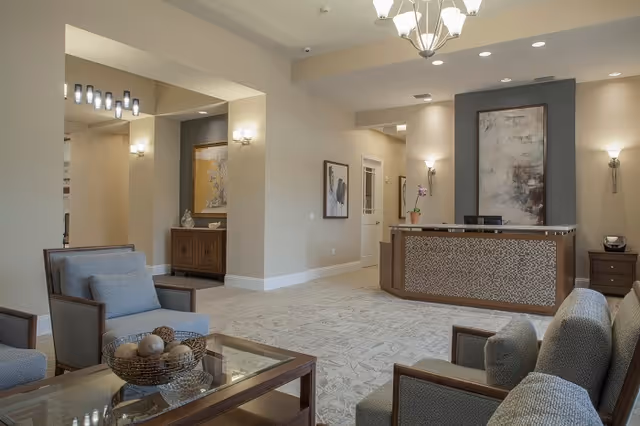 A spacious and well-lit reception area with a modern front desk featuring a mosaic tile design. The room has beige walls, carpeted floors with a subtle pattern, and comfortable seating including armchairs and a sofa around a glass coffee table with decorative spheres. Wall sconces and a chandelier provide warm lighting, and framed artwork decorates the walls.