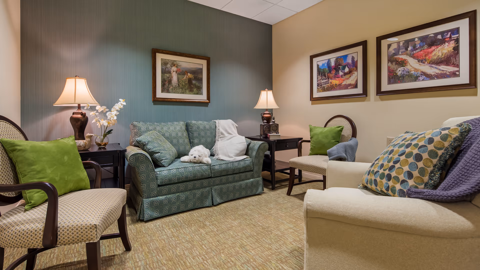 A cozy living room area in an assisted living facility with a green patterned loveseat, two armchairs with green and patterned cushions, two side tables with lamps, and framed artwork on the walls. A small white dog is resting on the loveseat.