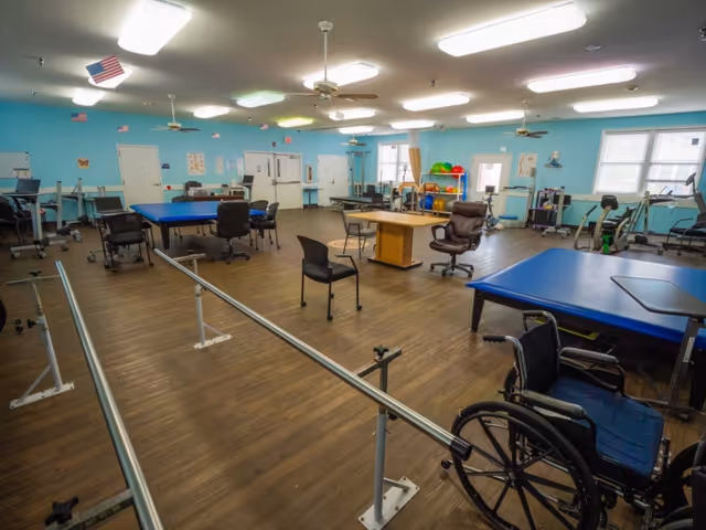 A spacious rehabilitation room with light blue walls and wooden flooring. The room contains parallel bars for walking exercises, several chairs including a wheelchair, therapy tables, exercise balls, and various physical therapy equipment. Ceiling fans and fluorescent lights are mounted on the ceiling, and windows allow natural light into the room.