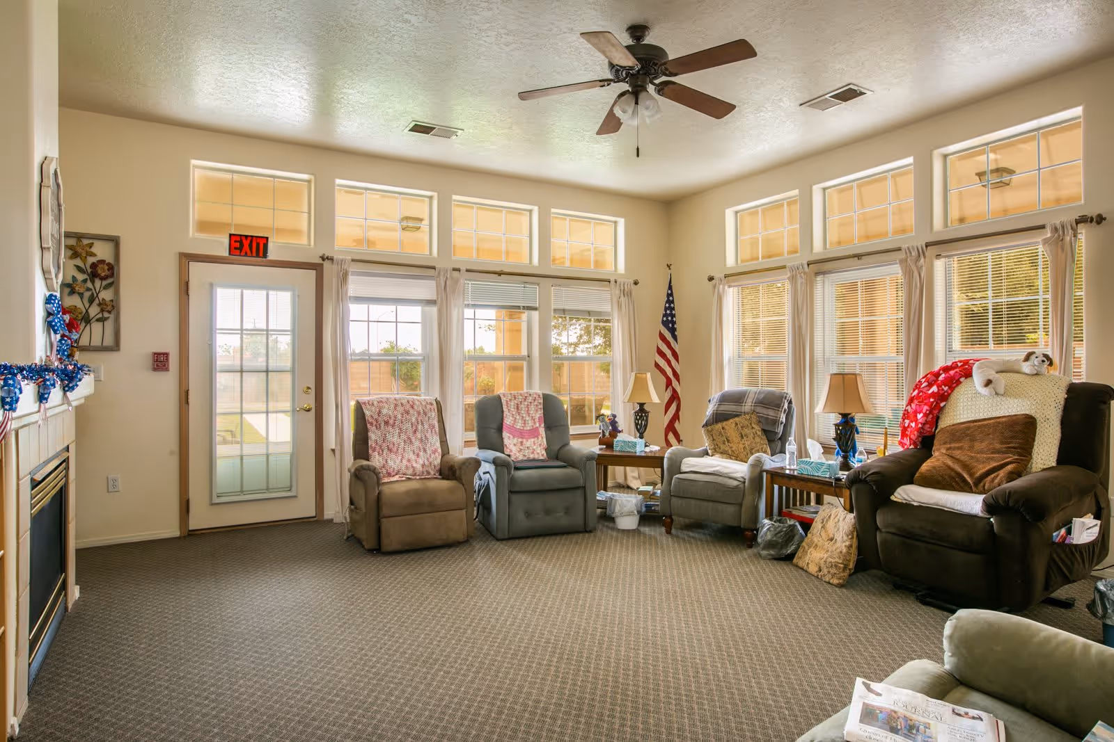 A bright and cozy living room with multiple recliner chairs arranged around a small wooden table. The room features large windows with beige curtains, a ceiling fan, a fireplace decorated with blue garland, and an American flag in the corner. The carpet is patterned and the walls are painted light beige.