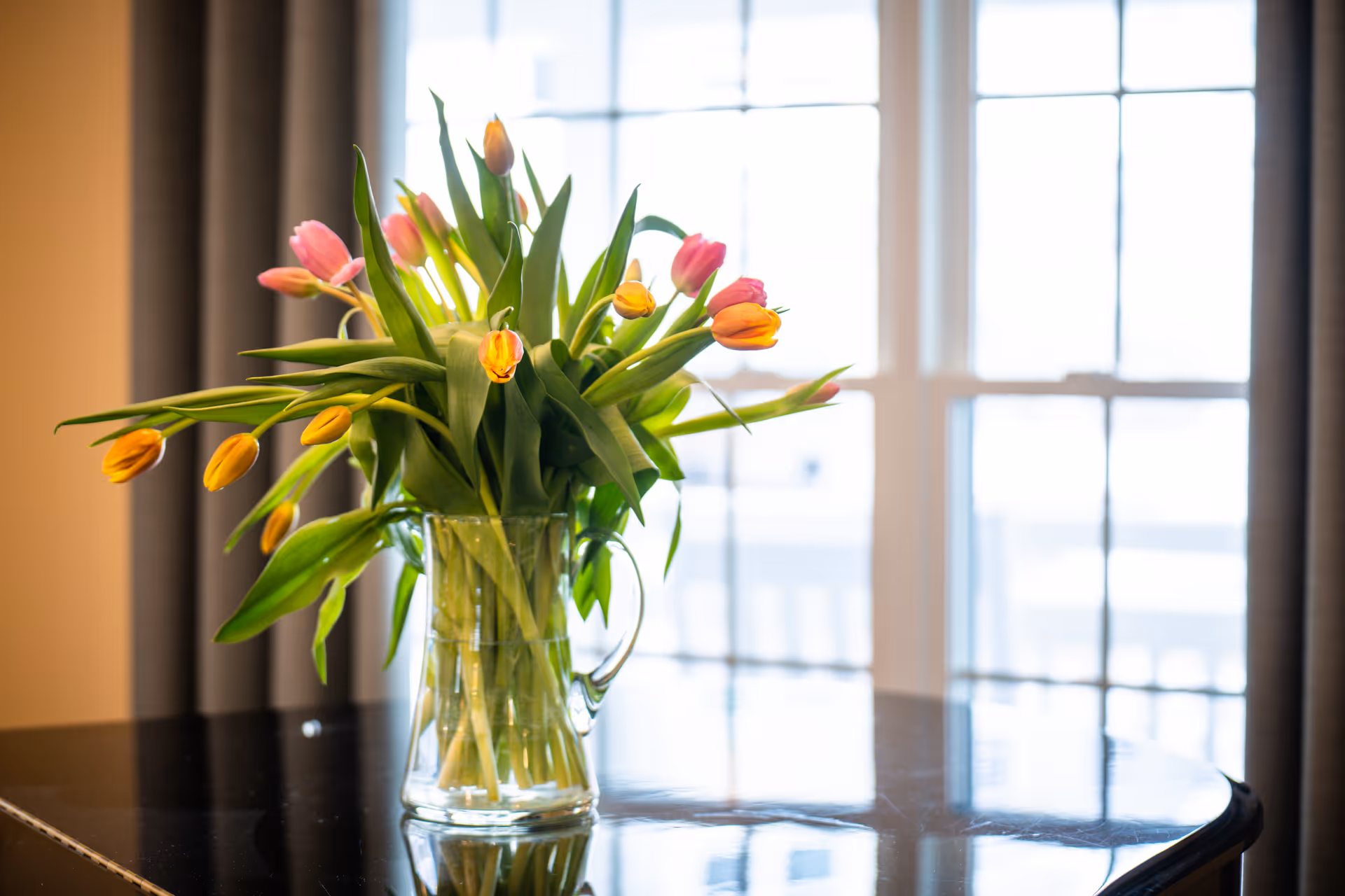 A glass pitcher filled with yellow and pink tulips sits on a glossy table in front of a bright window.