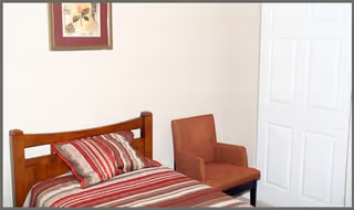 A small bedroom with a single wooden bed featuring striped bedding in shades of red, white, and brown. Next to the bed is a cushioned armchair in a reddish-brown color. A white closet door is visible on the right side of the image, and a framed floral picture hangs on the wall above the bed.