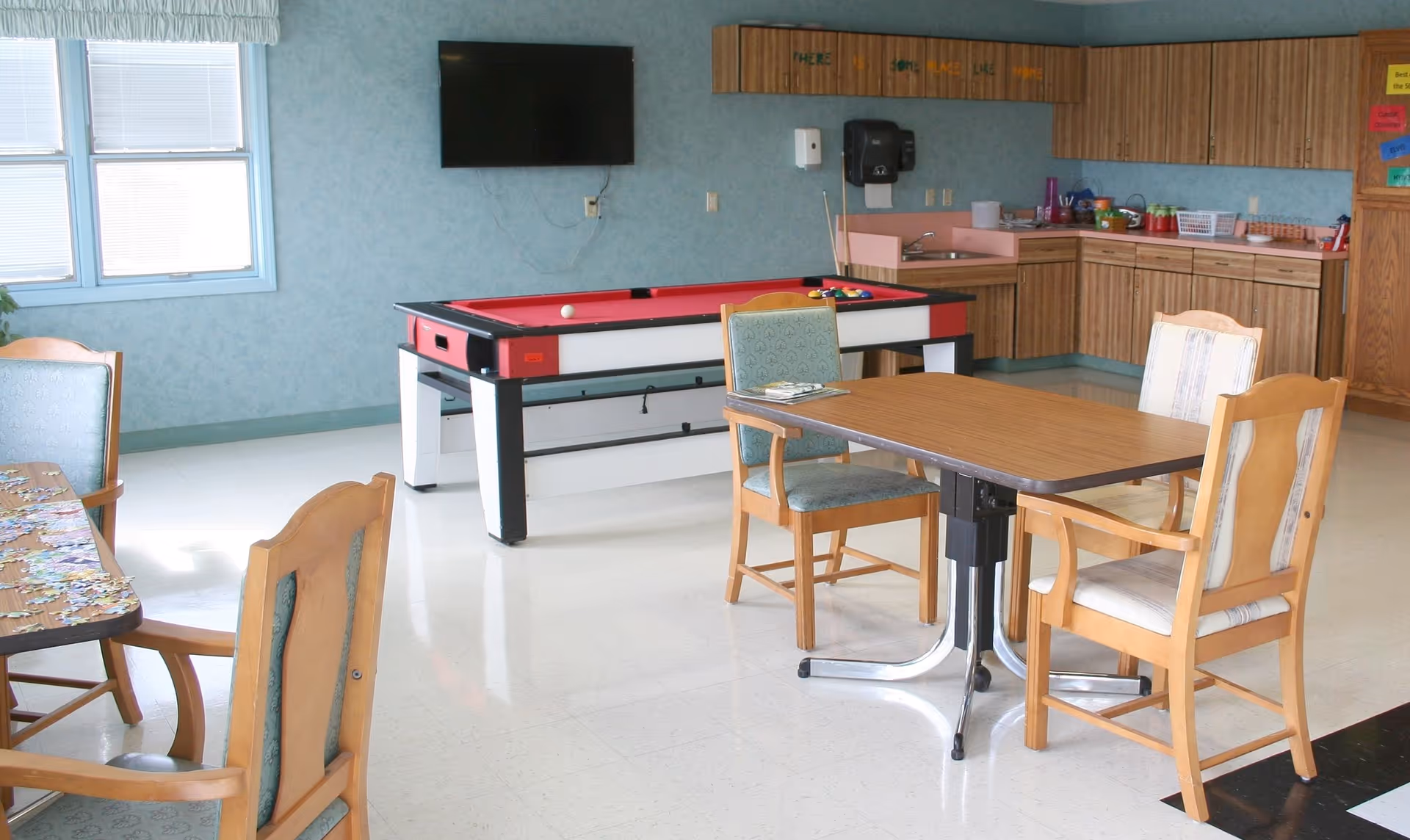 Bright common activity room with tables and chairs, a red pool table, wall-mounted TV, and a kitchenette along the back wall.