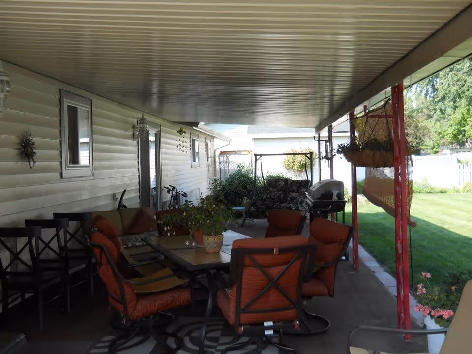Covered outdoor patio area with a table and several cushioned chairs, potted plants on the table and hanging, a barbecue grill, and a swing bench. The patio is adjacent to a house with white siding and windows, and there is a green lawn and garden visible beyond the patio.