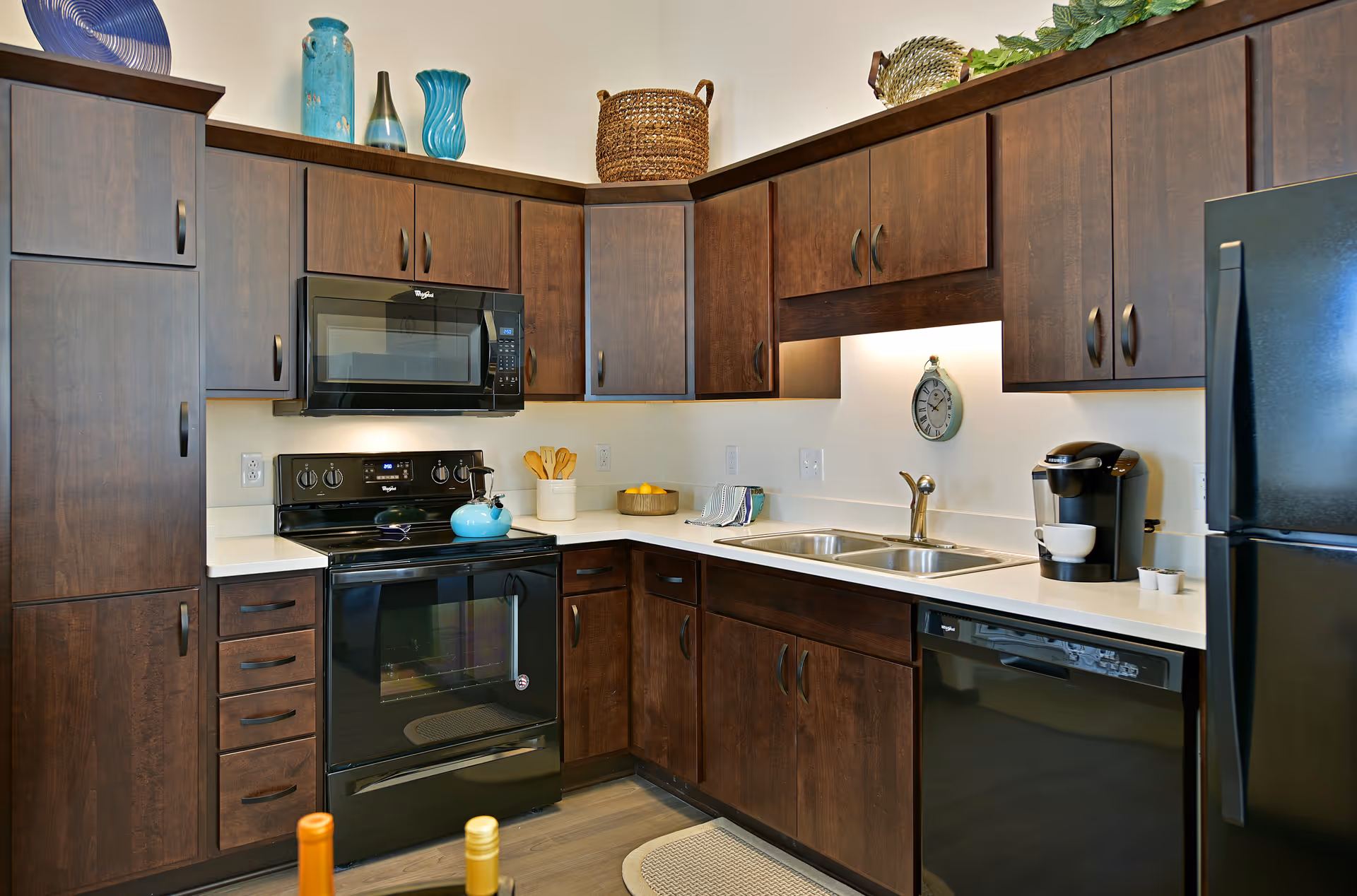 Modern kitchen with dark wood cabinets, black appliances including a stove, microwave, dishwasher, and refrigerator. The countertop has a double sink, a coffee maker with a cup, and some decorative items. There are decorative vases and a basket on top of the cabinets.